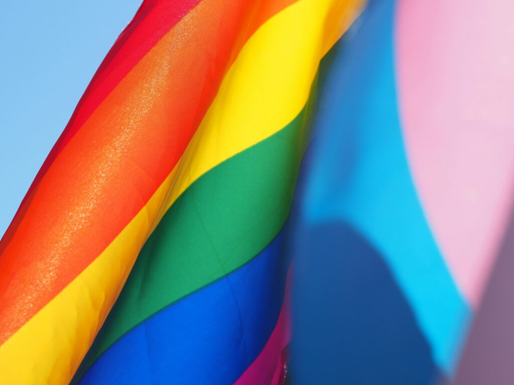 Närbild på två flaggor: en regnbågsfärgad Pride-flagga med röda, orange, gula, gröna, blå och violetta ränder och en pastellblå, rosa och vit transgender Pride-flagga, som båda vajar mot en klar himmel.