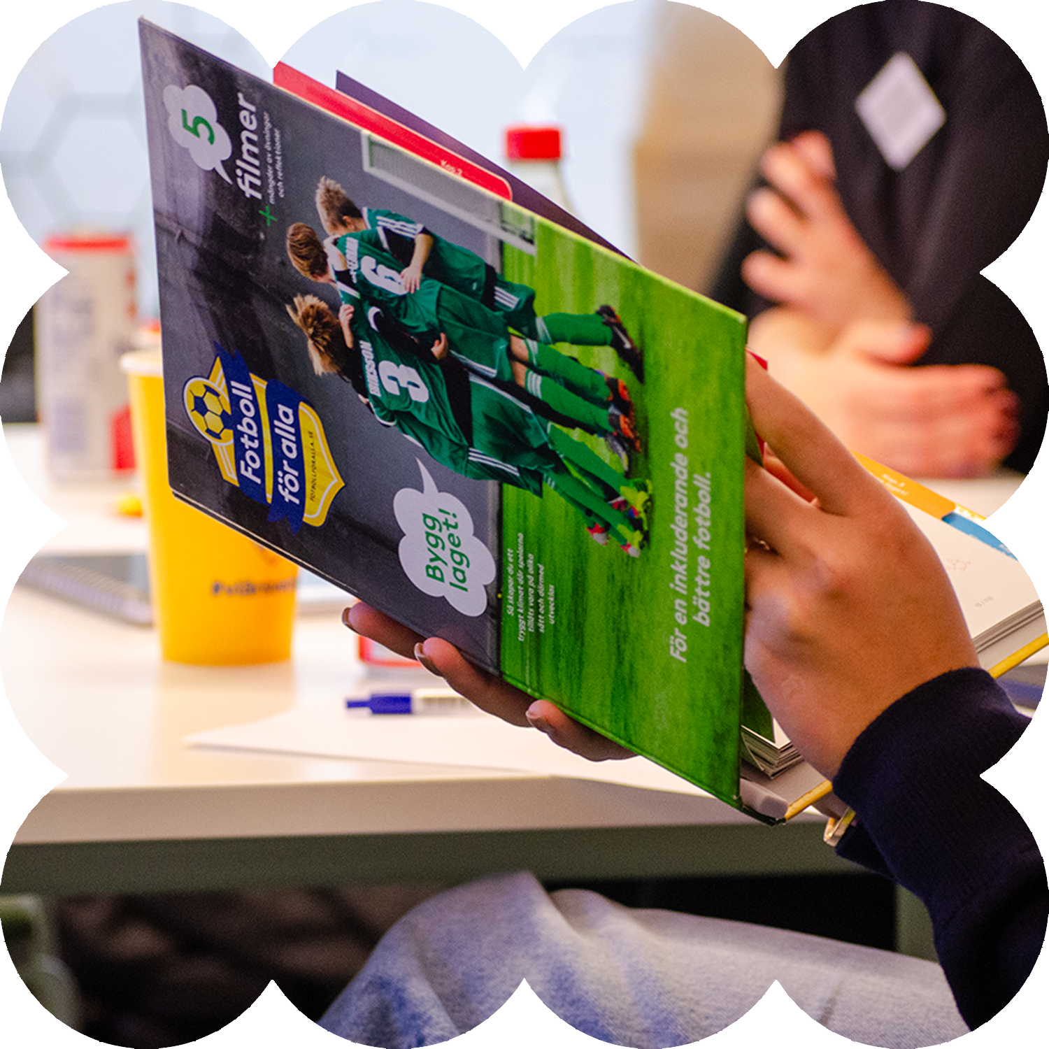 En person håller i en färgglad tidning med en grupp fotbollsspelare i gröna uniformer på omslaget, medan han sitter vid ett bord med papper, pennor och burkar utspridda.