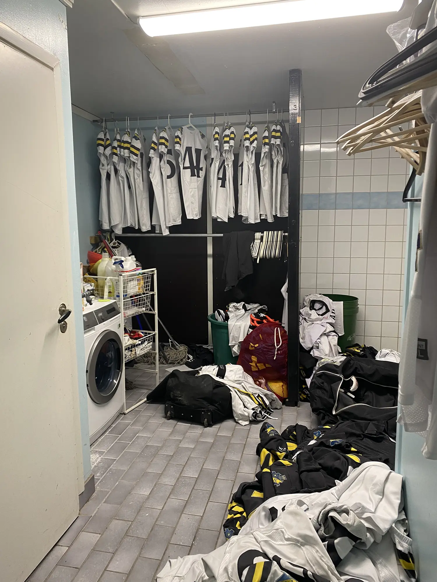 En tvättstuga med sporttröjor som hänger på tork, en tvättmaskin, hyllor med rengöringsmedel och högar med kläder och påsar utspridda på klinkergolvet.