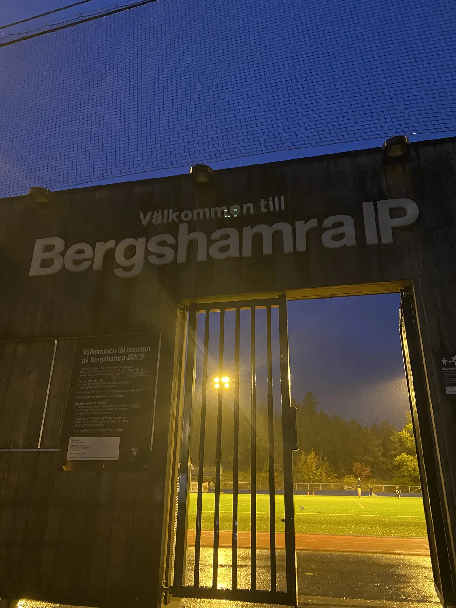 En metallgrind under en skylt med texten Välkommen till Bergshamra IP öppnar sig mot en starkt upplyst idrottsplats i skymningen, med mörka träd och en molnig blå kvällshimmel i bakgrunden.