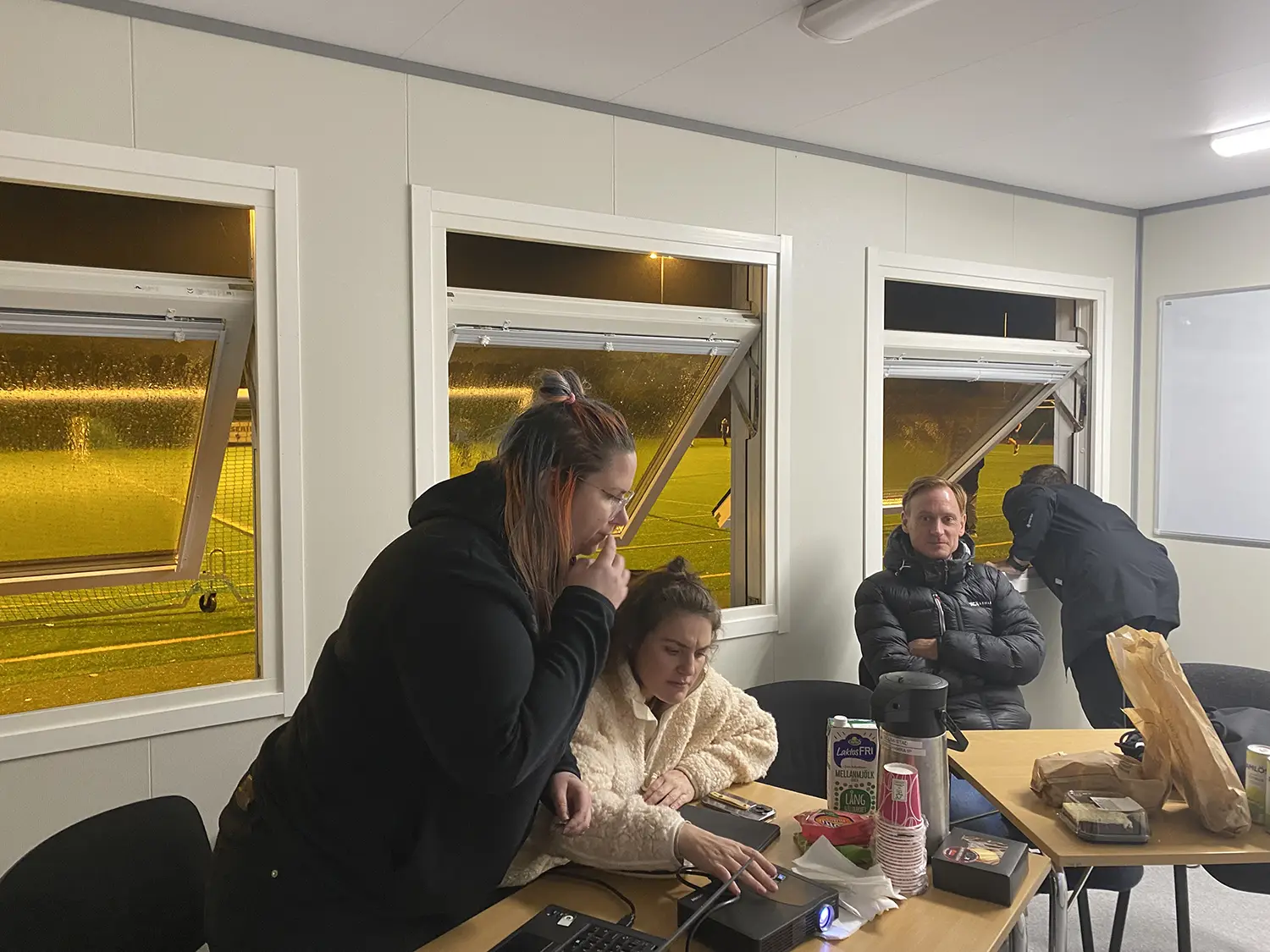 Fyra personer befinner sig i ett rum med öppna fönster på natten. Två kvinnor är fokuserade på en bärbar dator vid ett bord med snacks och kaffe. En man sitter i närheten och en annan står vid fönstret och tittar ut.