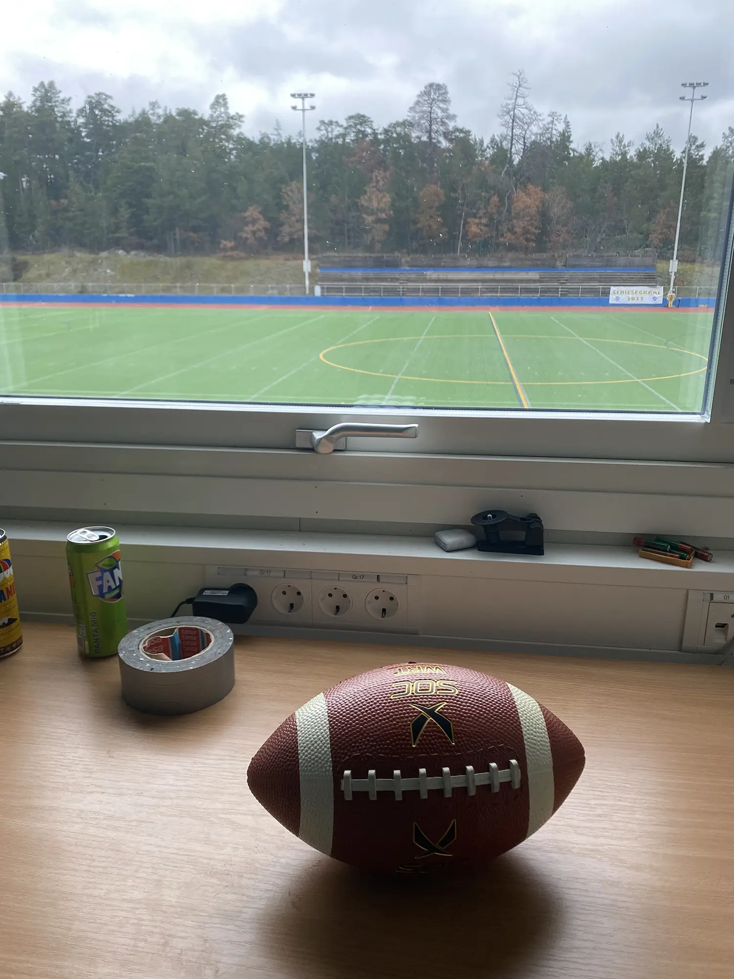 En fotboll ligger på ett skrivbord vid ett fönster med utsikt över en tom idrottsplats utomhus. På skrivbordet finns en burk Fanta, silvertejp, en pennvässare och andra pappersvaror. Träd och en molnig himmel syns utanför.