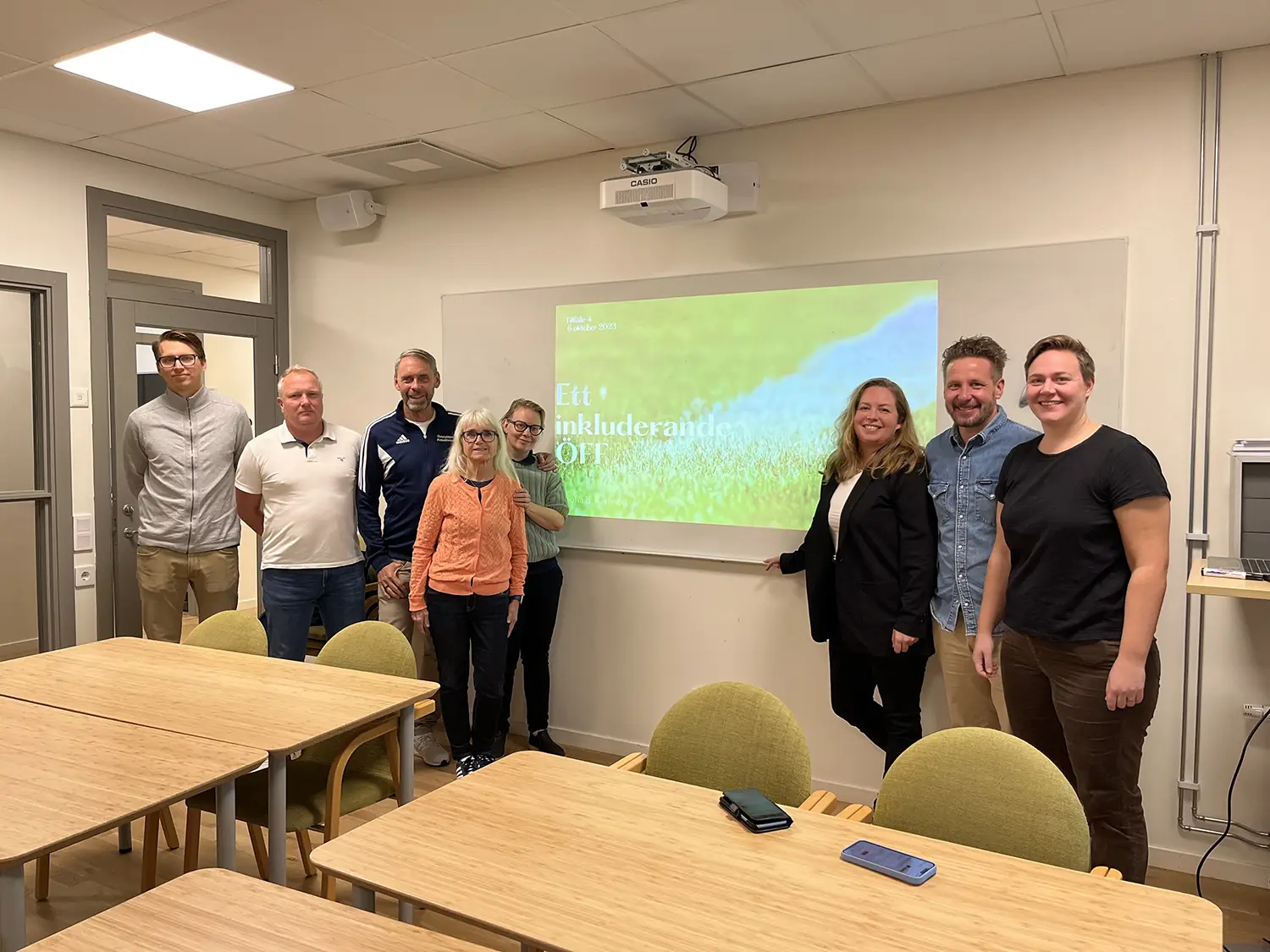 Åtta personer står och ler i ett klassrum intill en skärm som visar en presentation med titeln "Ett inkluderande ÖFT". Det finns bord och stolar i förgrunden och fluorescerande belysning ovanför.