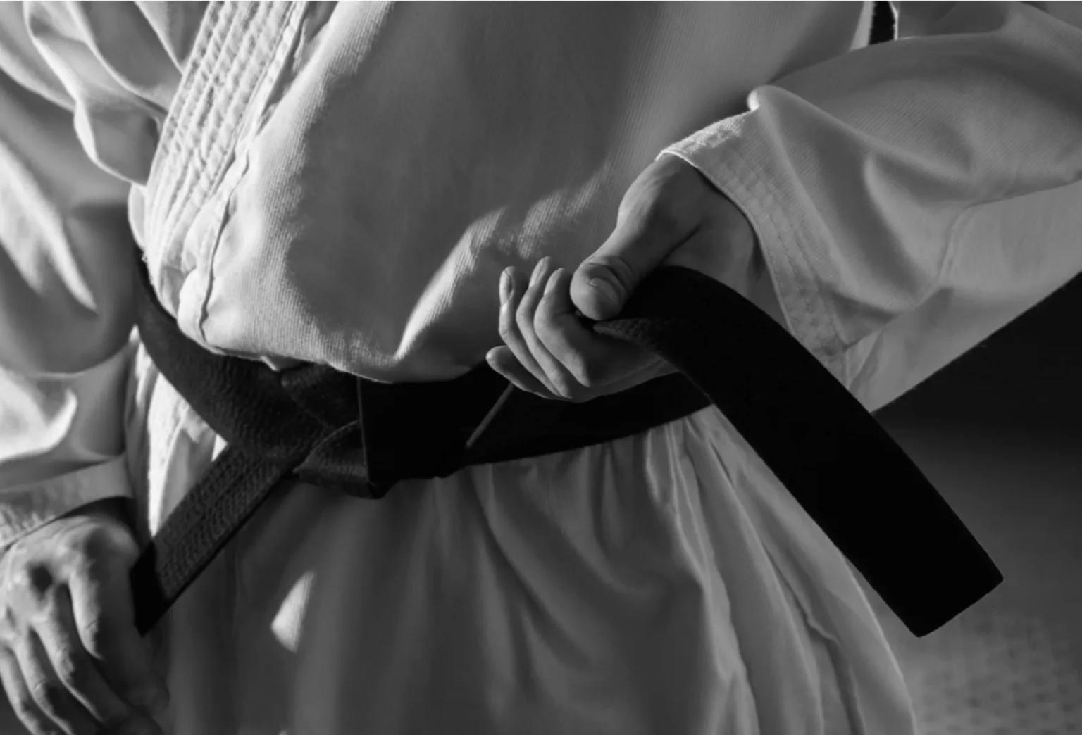 En person i kampsportsuniform knyter ett svart bälte runt midjan, bilden är i svartvitt.