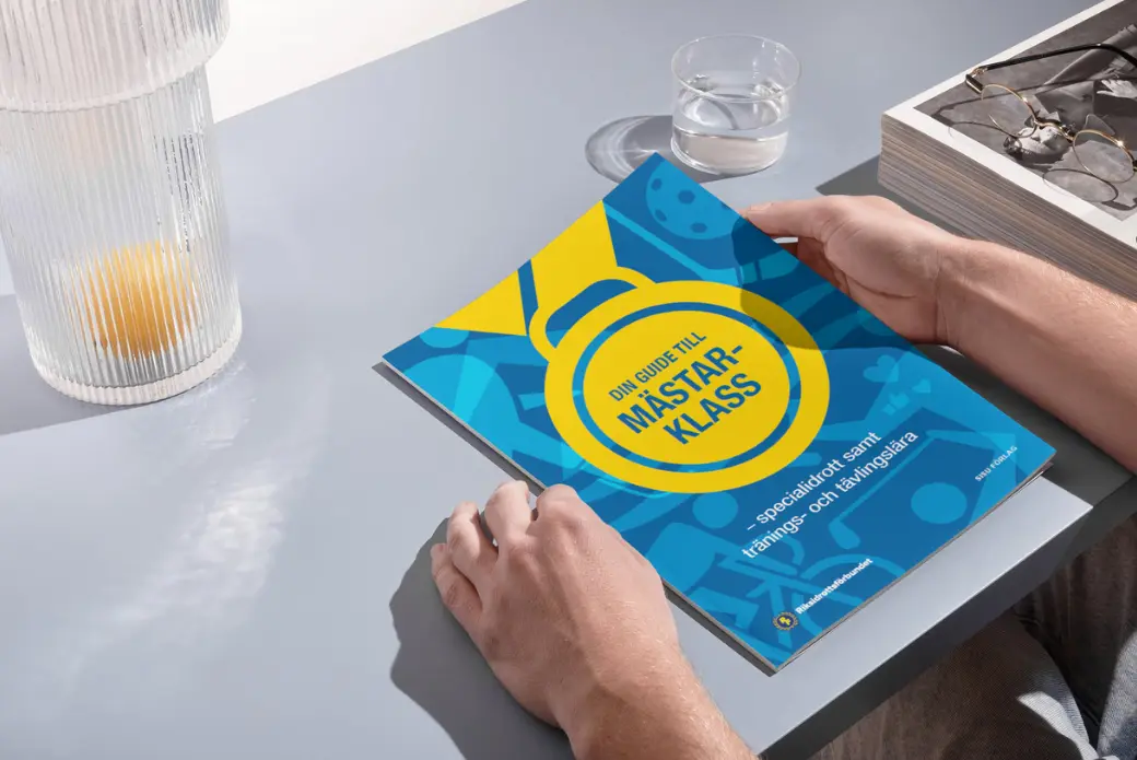 En person håller i en blågul broschyr med titeln Din guide till mästarklass på ett grått bord med ett glas vatten, en lampa och en bricka med glasögon bredvid.