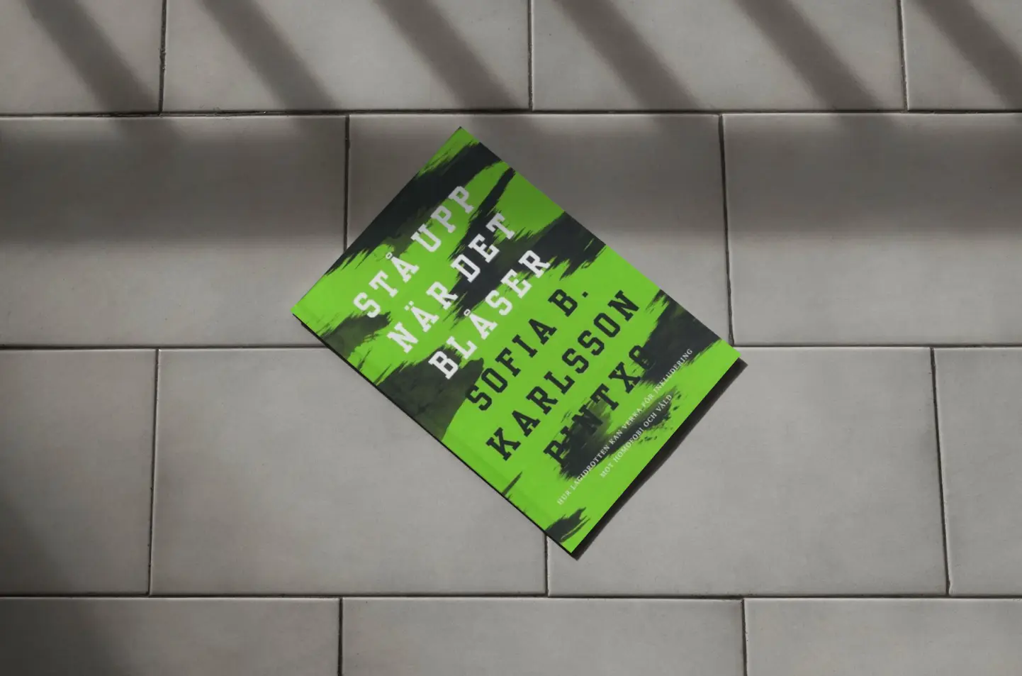 En grön och svart bok med titeln Stäupp när det blåser av Sofia B. Karlsson ligger på ljusgrå golvplattor, med skuggor från fönstergaller som kastas över scenen.