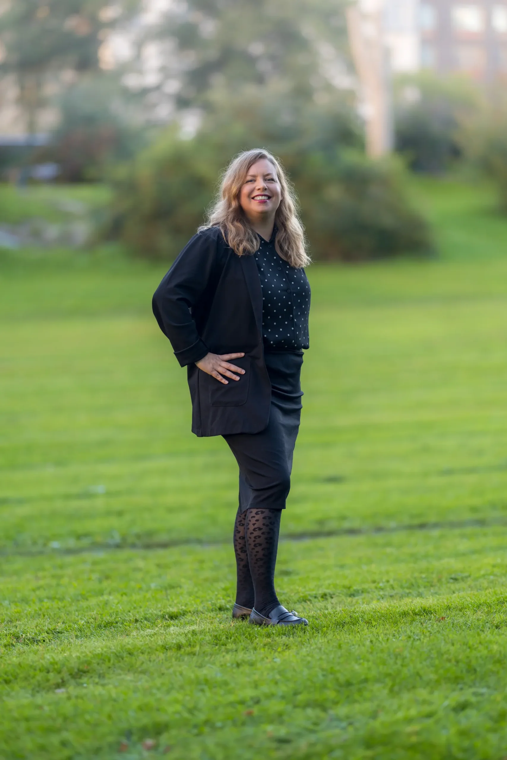 Sofia B Karlsson En kvinna med långt, vågigt blont hår står och ler på grönt gräs i en park, klädd i svart kavaj, prickig blus, svart kjol, mönstrade strumpbyxor och svarta skor. Träd och buskar är suddiga i bakgrunden.