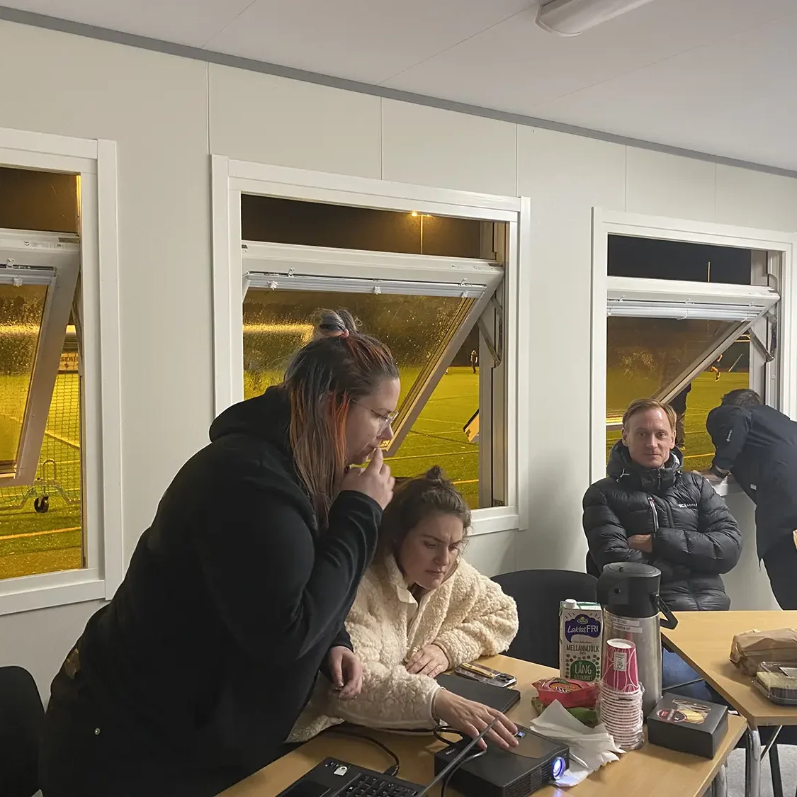 AIK Fyra personer befinner sig i ett rum med öppna fönster på natten. Två kvinnor är fokuserade på en bärbar dator vid ett bord med snacks och kaffe. En man sitter i närheten och en annan står vid fönstret och tittar ut.